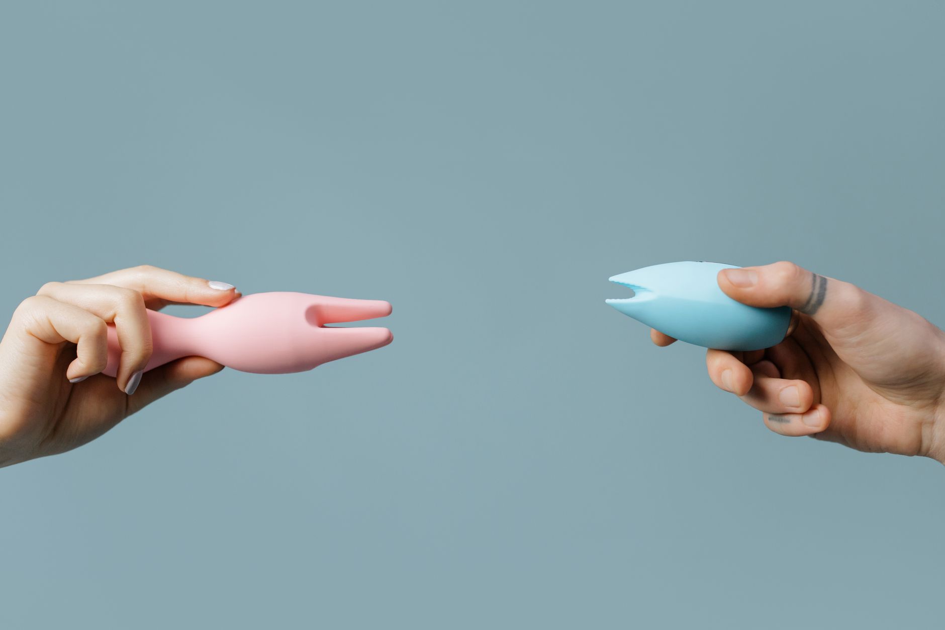 Two hands holding pink and blue silicone vibrators against a pastel background, representing intimacy and connection.