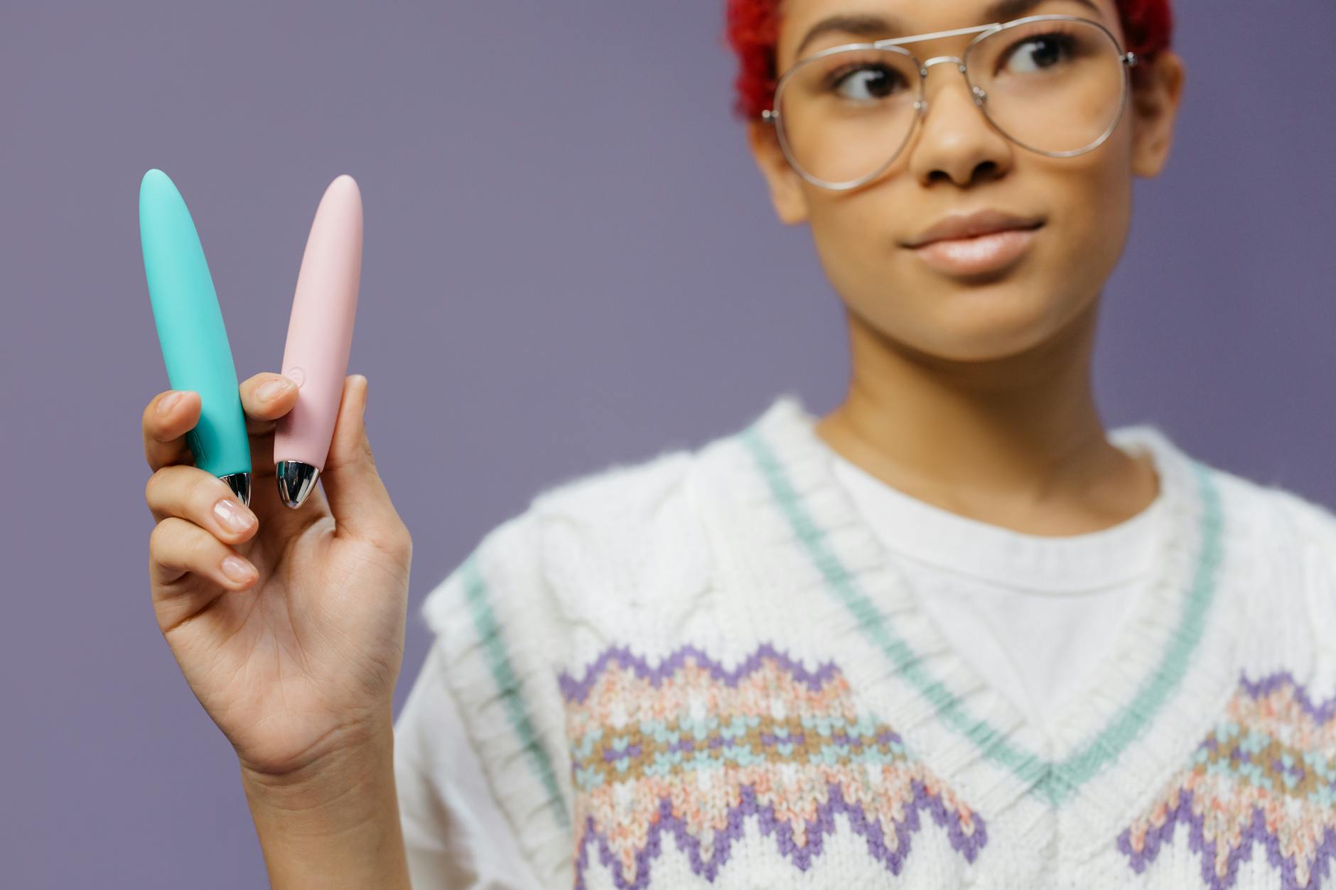 A woman with glasses holding colorful vibrators against a purple background, representing sexual wellness and confidence