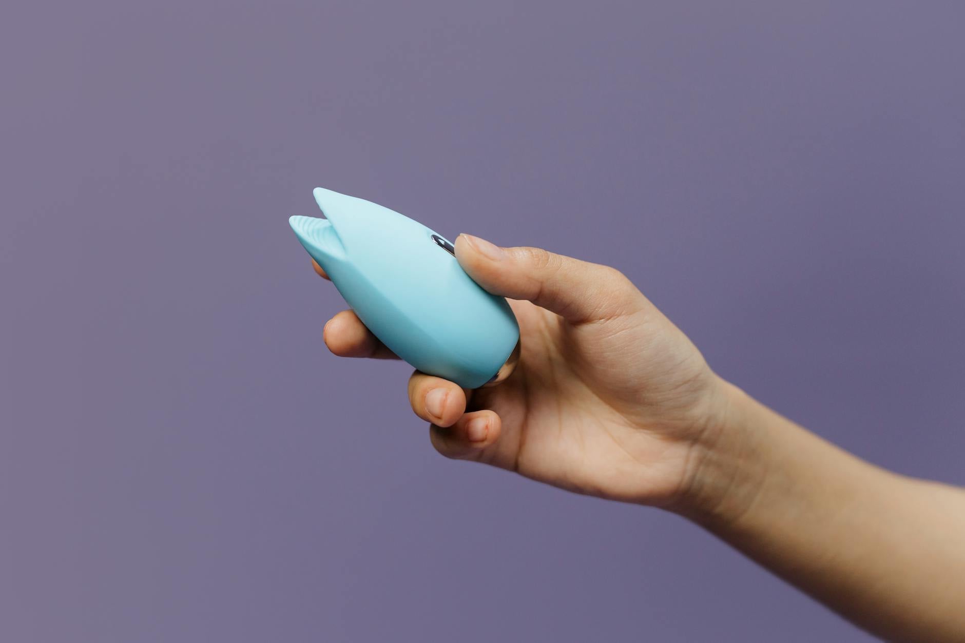 A hand holding a blue clitoral vibrator against a purple background, symbolizing self-exploration and pleasure